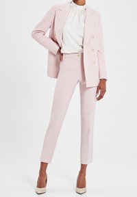 Blazer cintré rose clair avec des boutons dorés, associé à un pantalon ajusté assorti et un haut blanc à col montant. Tissu lisse, design épuré.