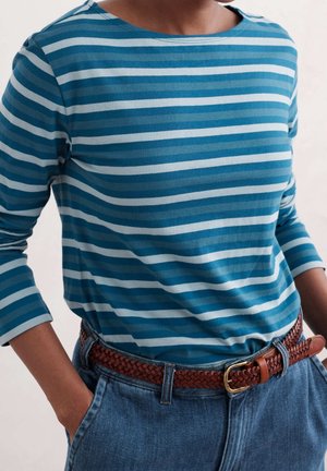 Osoba w niebiesko-białej pasiastym, długim rękawie koszulce wpuszczonej do niebieskich dżinsów z brązowym plecionym skórzanym paskiem, z rękami w kieszeniach.