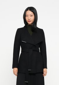 Mujer con cabello lacio y negro, vistiendo un abrigo negro con cinturón sobre una blusa verde oscura y una falda plisada negra, de pie contra un fondo blanco.