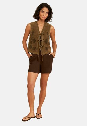 Frau trägt ärmellose braune Wildlederweste mit Blumenstickerei und Bändern, dunkelbraune Shorts und schwarze Sandalen, steht mit den Händen in den Taschen.