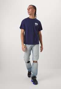 Navy blue cotton t-shirt with white graphic print. Worn with light blue distressed jeans featuring rips, and black and blue athletic shoes.