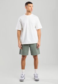 Valkoinen puuvillapaita lyhyillä hihoilla, yhdistettynä vihreisiin shortseihin, seisoo neutraalilla taustalla. Etenkin urheilukengät, joissa on yksityiskohtia.