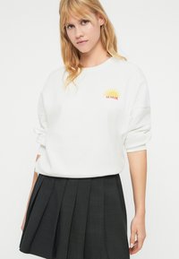 Jeune femme aux cheveux blonds portant un sweat-shirt blanc avec un soleil jaune et le texte "LE SOLEIL", assorti à une jupe plissée noire.
