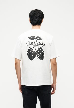 Mann sett bakfra iført hvit T-skjorte med svart terning- og bladdesign og teksten "Las Vegas Replay" på baksiden, svarte bukser.