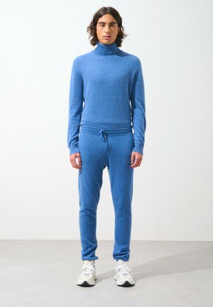 Blauwe gebreide coltrui en bijpassende joggers in een zachte, gestructureerde stof. Voorzien van een koordsluiting in de taille en aansluitende manchetten, gecombineerd met witte sneakers.