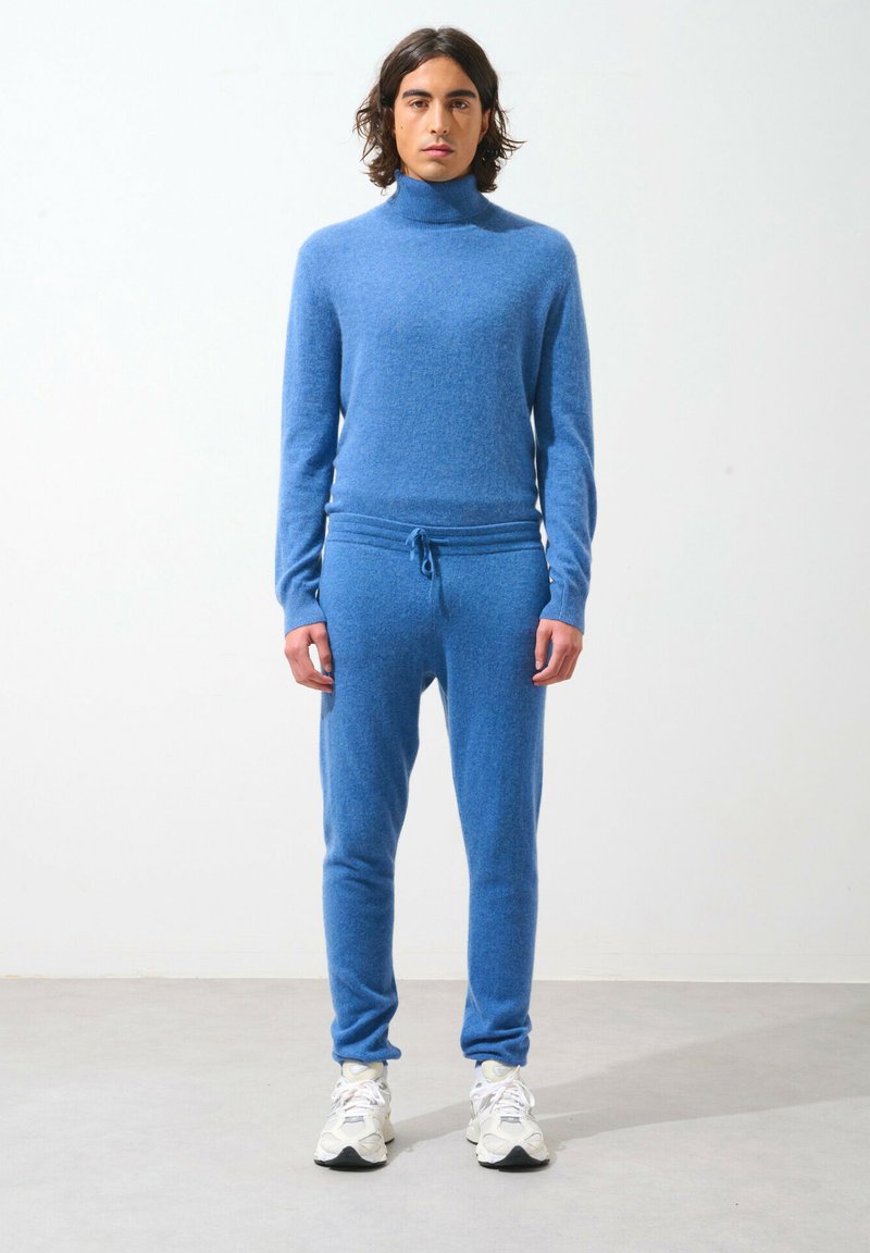 Blå stickad polo och matchande joggers i ett mjukt, strukturerat tyg. Har dragsko i midjan och åtsittande muddar, tillsammans med vita sneakers.