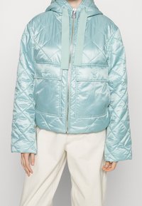 Helle blaue gesteppte Pufferjacke mit Kapuze, ausgestattet mit einem Frontreißverschluss, zwei großen Taschen und strukturierter Oberfläche.