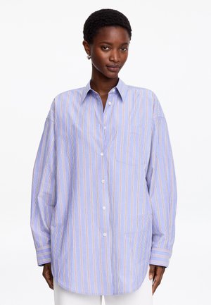 Mujer negra con una camisa azul claro de botones y corte holgado con rayas verticales blancas y beige, de pie frente a un fondo blanco.