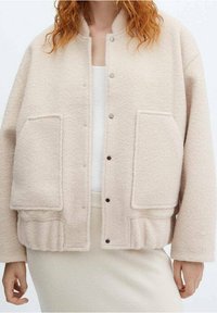 Light beige fleece jacket with a round collar, snap buttons, two front pockets, and a ribbed hem. Soft texture, relaxed fit.