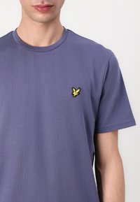 Lila kortärmad t-shirt i bomull, med en liten gul broderad fågel-logotyp på vänster bröstområde. Slät textur.