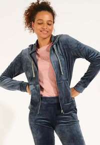 Felpa blu navy in velluto con zip e cappuccio, tasche frontali e dettagli argentati, abbinata a una maglietta rosa chiaro. Texture liscia.