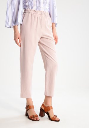 Frau trägt hellrosa, elastische, gekürzte Hosen mit Bündchen und braune, lederne Sandalen mit Absatz und Schnallen, steht vor weißem Hintergrund.