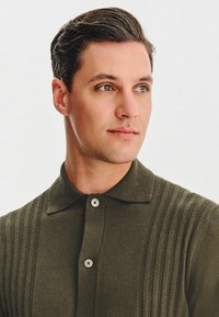 Ciemnozielona koszulka polo z teksturowanym wzorem w paski, z kołnierzykiem w kształcie szpicu i dwoma guzikami z przodu. Miękki materiał i dopasowany krój.