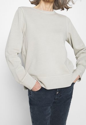 Kobieta ubrana w jasnobeżową, długorękawową bluzę i ciemnoniebieskie dżinsy, stojąca z rękami w kieszeniach na białym tle.