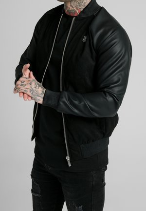 Personne aux mains et au cou tatoués portant une veste noire zippée avec des manches en cuir et un jean noir usé sur un fond uni.