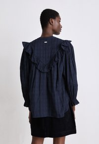 Marineblaue Bluse mit karierter Textur, ausgestattet mit einem rüschierten V-Ausschnitt und langen Puffärmeln. Die Rückansicht zeigt ein minimalistisches Design.