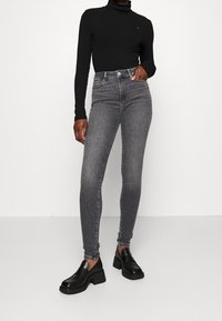 Jeans de denim gris de cintura alta con corte slim, combinados con un suéter de cuello alto negro de canalé y zapatos negros de tacón grueso. Diseño minimalista.