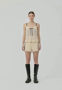 Woman standing wearing beige lace camisole and matching shorts with black trim, paired with knee-high black boots, neutral background.