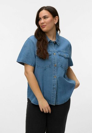 Mujer con cabello largo y castaño, vestida con una camisa de mezclilla de manga corta abotonada y pantalones negros, de pie con una mano en el bolsillo.