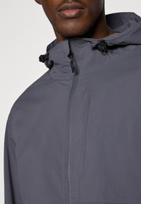 Giacca leggera grigia con colletto alto, chiusura a zip e cappuccio regolabile con cordino. Tessuto liscio con minimi dettagli.