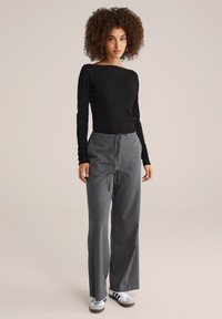 Vrouw met krullend haar, gekleed in een zwarte lange mouw top, grijze wijde broek met trekkoord en witte sneakers met zwarte strepen, staand.