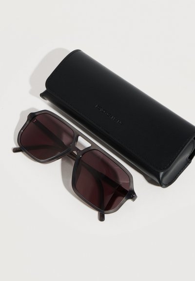 Μαύρα τετράγωνα γυαλιά ηλίου με σκούρους φακούς δίπλα σε μαύρη ορθογώνια δερμάτινη θήκη Dolce & Gabbana πάνω σε λευκή επιφάνεια.