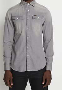 Camicia in denim grigio con colletto a punta, due tasche sul petto e bottoni a pressione argentati. Presenta una texture leggermente sbiadita e un orlo curvo.