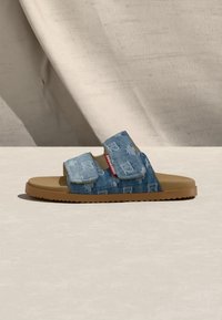 Sandale à glissière en denim à deux brides avec motif usé et semelle marron, posée sur une surface claire devant un fond en tissu beige.