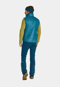 Tessellierte, teal-farbene, isolierte Weste mit hohem Kragen, kombiniert mit einem grünen Langarmoberteil und blauen Hosen. Das Outfit umfasst strapazierfähige Stiefel und strukturierte Akzente.