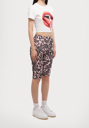 Femme portant un crop top blanc avec un motif de lèvres et de langue, un short longueur genou à imprimé léopard rose, des chaussettes blanches et des baskets blanches.