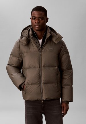 Homme portant une veste matelassée marron avec capuche, fermée par une fermeture éclair, une main dans la poche, sur un fond gris clair uni.