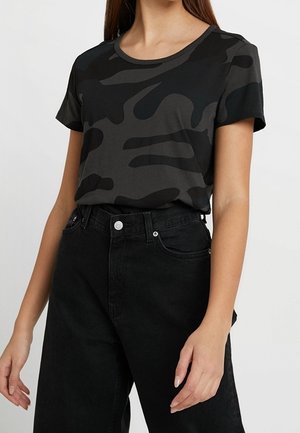 Woman wearing short-sleeve black and dark gray abstract camo-patterned t-shirt tucked into high-waisted black jeans.