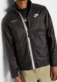 Nike Air takki mustana, jossa on vetoketjulista, tekstuuripinta ja valkoiset yksityiskohdat. Pitkät hihat elastisilla resoreilla. Kevyt kangas.