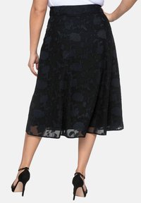 Mujer vestida con una falda midi negra con estampado floral y dobladillo transparente, de pie con una mano en la cadera y tacones altos negros con tiras al tobillo.
