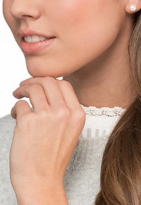 Orecchino a perla a bottone, tono di pelle chiaro, mano morbida appoggiata sul collo, maglione grigio testurizzato con bordo in pizzo bianco, capelli lisci che cascano.