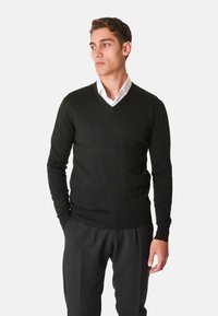 Maglione nero a maglia con scollo a V, maniche lunghe e polsini a coste. Indossato sopra una camicia bianca con colletto, abbinato a pantaloni scuri.