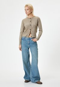 Cardigan beige lavorato a maglia con bottoni, orlo a coste e maniche lunghe, abbinato a jeans blu a gamba larga. Tessuto casual e strutturato.