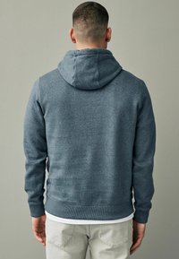 Sudadera azul de mezcla de algodón, con capucha con cordón, puños y dobladillo acanalados. Llevada con pantalones de color claro, vista desde atrás.