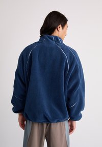 Navy blue fleece pullover with a high collar, loose fit, white stitching accents, and textured surface; worn over light beige pants.