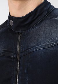 Närbild av en mörk denimjacka med hög krage som är stängd med tryckknapp och en synlig framtida dragkedja, bärs av en person.