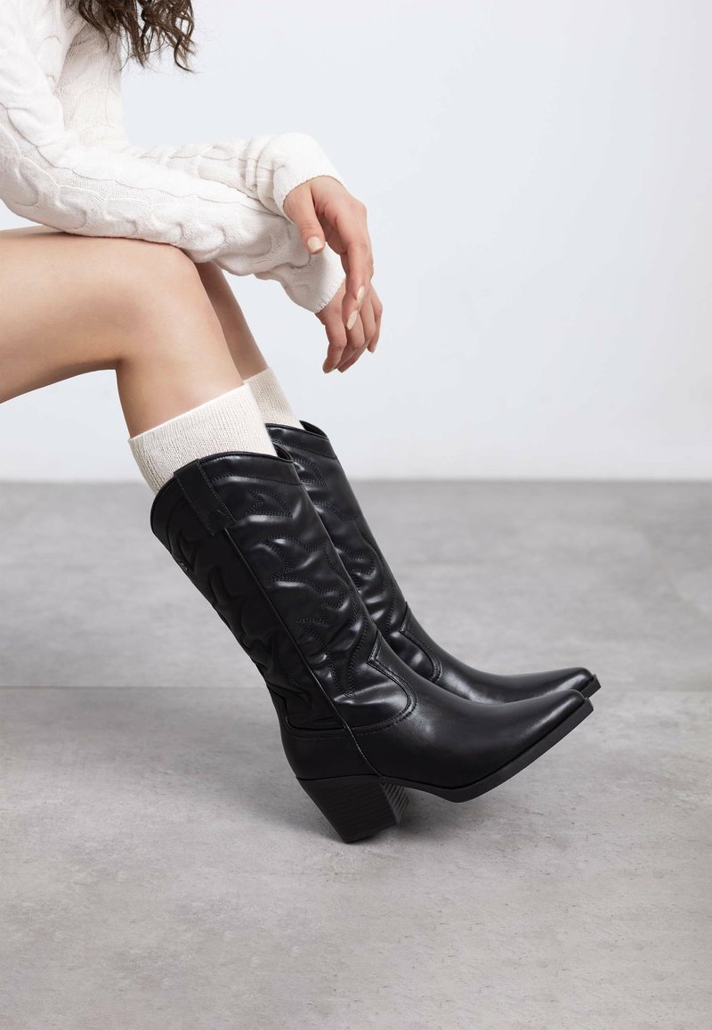 Botas de vaquero de cuero negro con punta afilada, detalles texturizados y tacón apilado, combinadas con calcetines blancos con relieve.