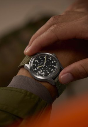 Ruka upravuje hodinky Timex s černým ciferníkem a zeleným látkovým páskem na zápěstí s tmavou pokožkou, oblečená v zelené bundě a šedém manžetu.