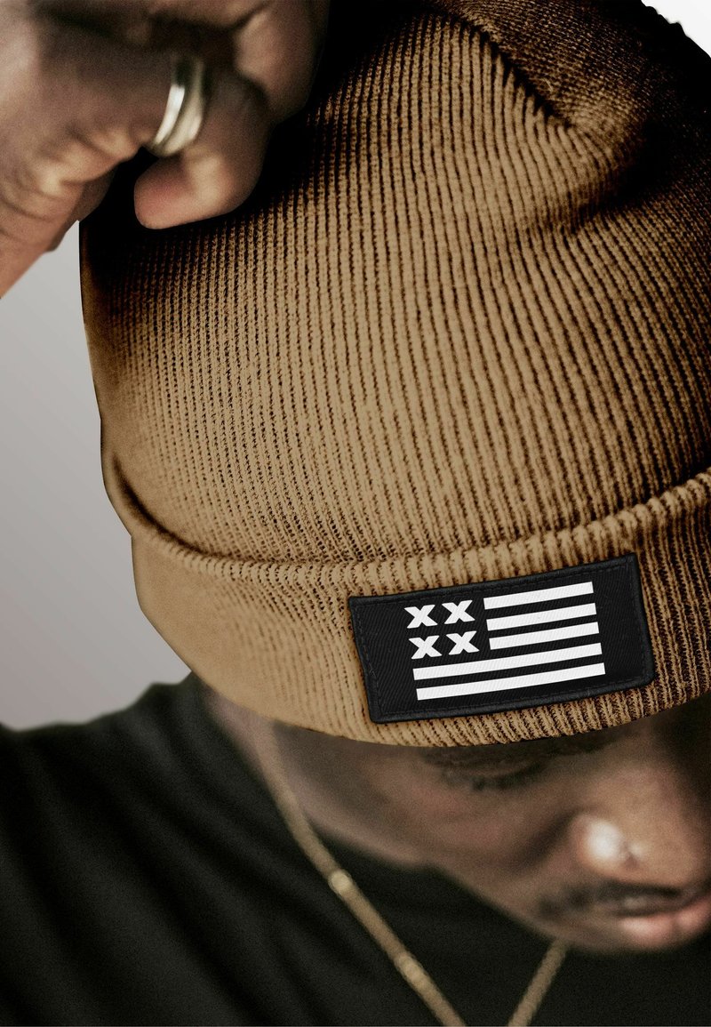 Brown ribbed beanie hat featuring a black patch with white stripes and symbols. It has a soft texture and a snug design for warmth.