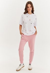 Biele tričko s krátkym rukávom, s farebnými vyšívanými vzormi, v kombinácii s ružovými teplákmi, ktoré majú logo a bočné písanie. Hnedo-biele tenisky.