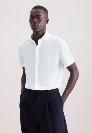 Uomo che indossa una camicia bianca fresca a maniche corte con bottoni e pantaloni plissettati blu scuro, in piedi con le mani nelle tasche.