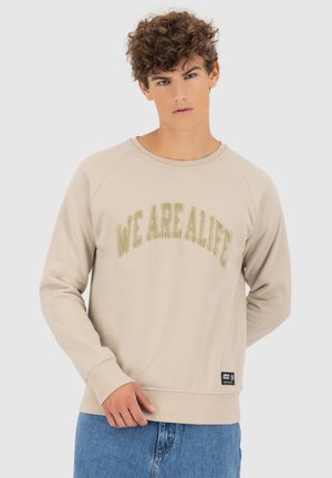 Jonge man met krullend haar, gekleed in een beige sweatshirt met de tekst "WE ARE ALIFE" en blauwe jeans, poseert tegen een effen lichte achtergrond.