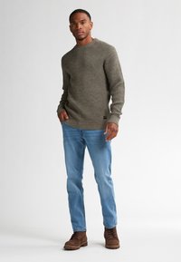 Olijfgroene gebreide trui met een gestructureerd patroon, geribbelde manchetten, gecombineerd met lichtblauwe jeans en bruine veterschoenen.