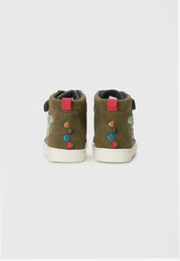 Bottines en suede vert olive avec des semelles en caoutchouc blanc, des accents brodés et des onglets colorés en rouge, orange et bleu de chaque côté.