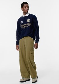 Marineblaues Langarm-Polohemd mit weißem Kragen, goldener Schriftzug "Community Culture", kombiniert mit khakifarbenen Cargo-Hosen und beigen Wanderschuhen.