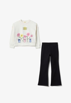 Felpa bianca corta con design floreale colorato e personaggi; abbinata a pantaloni neri a zampa, entrambi realizzati in tessuto morbido.